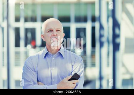 Un uomo d'affari di alto livello si trova fuori da un moderno edificio di uffici, la sua espressione è premurosa quando possiede uno smartphone, forse contemplando una strategia aziendale Foto Stock