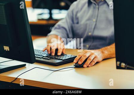 Il lavoratore esperto in una camicia blu si concentra sullo schermo di un computer in un ufficio, mostrando concentrazione e impegno nei suoi compiti Foto Stock