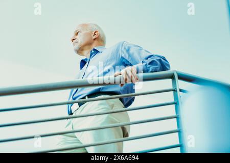 L'anziano con una camicia blu si appoggia contro una ringhiera, con un aspetto premuroso mentre guarda in lontananza, riflettendo sul passato e sul futuro della vita Foto Stock