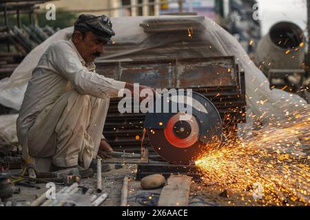 Il povero vecchio saldatore pakistano che taglia tubi e aste metalliche nella sua officina di strada con un taglierino elettrico, causando fiamme e scintille Foto Stock