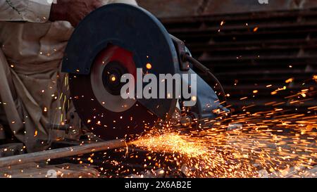 Saldatore che taglia tubi e aste metalliche nella sua officina stradale con un taglierino elettrico che provoca fiamme e scintille Foto Stock