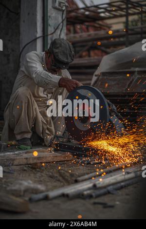 Il povero vecchio saldatore pakistano che taglia tubi e aste metalliche nella sua officina di strada con un taglierino elettrico, causando fiamme e scintille Foto Stock