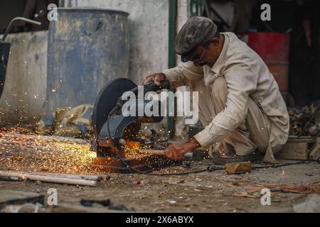 Il povero vecchio saldatore pakistano che taglia tubi e aste metalliche nella sua officina di strada con un taglierino elettrico, causando fiamme e scintille Foto Stock