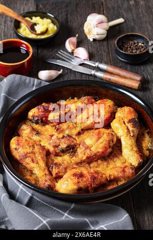 cosce di pollo cotte in salsa di ananas tritata in un piatto nero su un tavolo di legno rustico scuro con ingredienti e posate sullo sfondo, via verticale Foto Stock