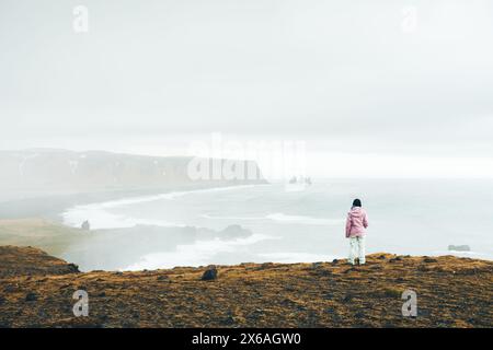 Primo piano donna caucasica in una fredda postazione contempla in riva al mare solo sul punto panoramico in una giornata ventosa e piovosa. Spiaggia islandese di sabbia nera, famosa per l'oceano atlantico Foto Stock