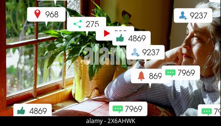 Immagine di icone dei social media su una donna anziana caucasica che guarda fuori dalla finestra a casa Foto Stock