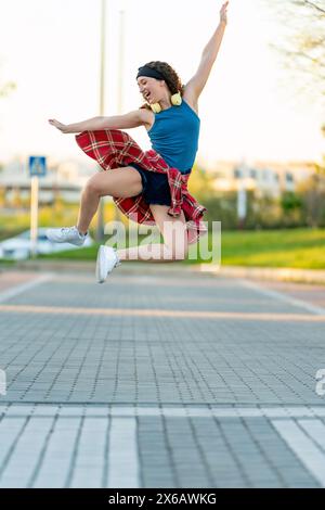 Giovane donna vivace con canotta blu e camicia a quadri salta in alto, ascoltando la musica attraverso le cuffie su un sentiero soleggiato del parco. Foto Stock