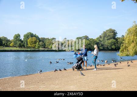 Hyde Park London, giovane coppia di trent'anni spinge il bambino in carrozzina accanto al lago Serpentine giorno d'autunno soleggiato, 2023 settembre, Londra, Regno Unito Foto Stock