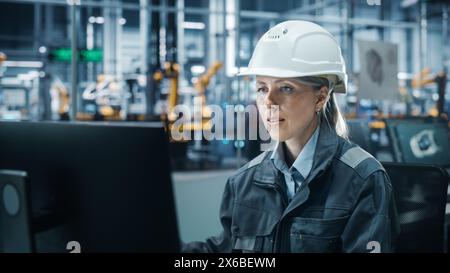 Ufficio della fabbrica di automobili: Ritratto di un ingegnere capo donna sicura di sé che indossa l'Hard Hat che lavora su un computer desktop. Tecnico della linea di assemblaggio del braccio robotico automatizzato che produce veicoli elettrici ad alta tecnologia Foto Stock