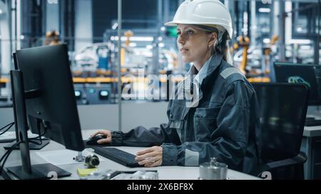 Ufficio della fabbrica di automobili: Ritratto di un ingegnere capo donna sicura di sé che indossa l'Hard Hat che lavora su un computer desktop. Tecnico della linea di assemblaggio del braccio robotico automatizzato che produce veicoli elettrici ad alta tecnologia Foto Stock
