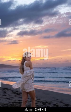 Una giovane donna avvolta da uno scialle bianco si erge su una spiaggia sabbiosa, che guarda le spalle sullo splendido cielo del tramonto con sfumature rosa e arancio Foto Stock