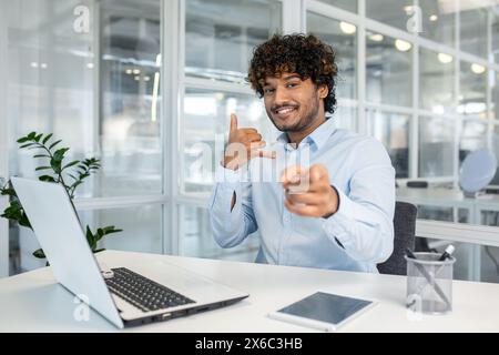 Allegro uomo d'affari con una camicia blu che fa un gesto chiamato e punta la fotocamera mentre è seduto in un moderno ufficio. Foto Stock