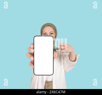 Donna musulmana caucasica in hijab che mostra smartphone. Immagine vuota del grande schermo bianco vuoto. Display gadget con dito puntato. Dimostrazione dello spazio libero. Foto Stock