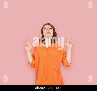 bella giovane ragazza caucasica dai capelli rossi che tiene le dita incrociate. Guardare in alto, esprimere un desiderio in attesa di un momento speciale. Indossa una camicia arancione. Foto Stock