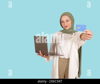 moderna ragazza musulmana caucasica in hijab che tiene aperto il laptop e mostra la carta di credito di debito. Guarda la fotocamera. Consigliare l'e-commerce. Foto Stock
