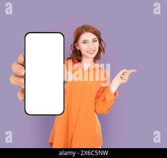 sorridente donna caucasica abbastanza felice che tiene in mano mostrando lo smartphone bianco vuoto dispositivo a schermo vuoto mock-up. Puntare il dito indice da un lato all'altro dello spazio di copia. Foto Stock