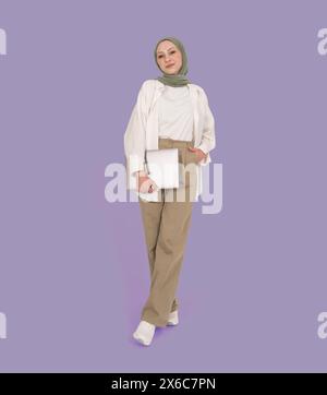 Vista frontale completa, giovane donna musulmana caucasica in hijab con laptop chiuso. Indossa pantaloni beige, camicia bianca e foulard verde. Foto Stock