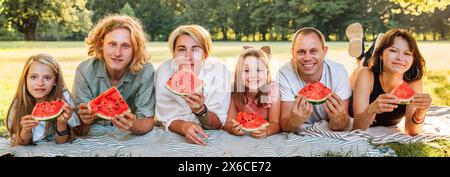 Una grande famiglia si stende sulla coperta da picnic nel parco cittadino sotto l'albero di tiglio durante il fine settimana domenica giorno di sole, sorridendo, ridendo alla macchina fotografica e mangiando l'acquavite Foto Stock