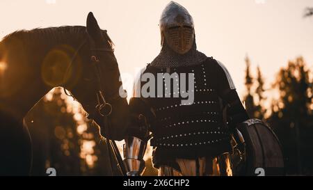 Cavaliere medievale che conduce il suo cavallo nella battaglia per combattere. Guerriero in armatura, casco, Spada e Stallion purosangue. Luci cinematografiche, guerra, invasione, conquista. Vista frontale verticale Foto Stock