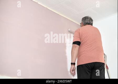 Un uomo anziano è visto rinnovare la sua casa, applicando un nuovo strato di vernice rosa su un muro con un rullo, catturato in un momento di concentrazione e auto Foto Stock