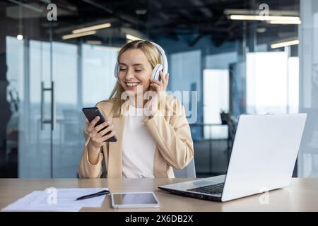 Una giovane donna d'affari felice ascolta la musica con le cuffie mentre utilizza lo smartphone e il computer portatile in un ambiente luminoso. Una piccola pausa durante il lavoro. Foto Stock