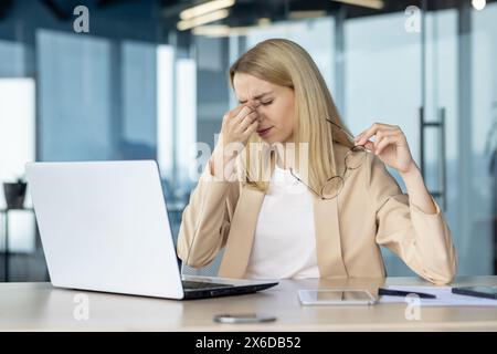 Una donna d'affari si sente stressata e affaticata mentre lavora al suo notebook in un ufficio moderno. Tiene gli occhiali e si strofina gli occhi a causa dell'esaurimento. Foto Stock