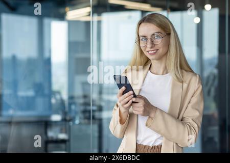 Giovane donna d'affari sicura che utilizza uno smartphone in un ufficio moderno. Sorride e indossa gli occhiali, ritraendo un aspetto amichevole e professionale. Foto Stock