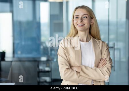 Ritratto di una giovane donna d'affari sicura di sé con le braccia incrociate, sorridente in un ufficio moderno illuminato dal sole. Trasuda professionalità e positività in un ambiente aziendale. Foto Stock