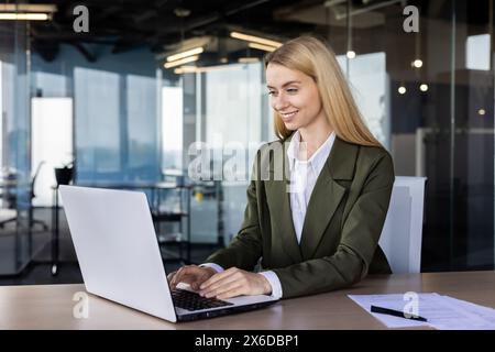 Giovane donna professionale sicura di sé in un blazer verde che utilizza un notebook in un ambiente d'ufficio moderno, che mostra efficienza e concentrazione. Foto Stock