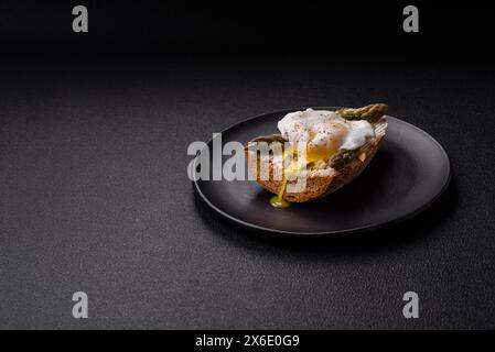 Colazione deliziosa e abbondante, con uova in camicia su pane tostato con formaggio spalmabile, asparagi, sale e spezie su sfondo di cemento scuro Foto Stock