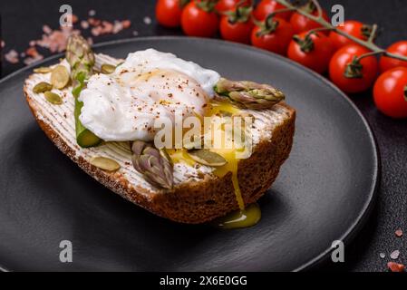 Colazione deliziosa e abbondante, con uova in camicia su pane tostato con formaggio spalmabile, asparagi, sale e spezie su sfondo di cemento scuro Foto Stock