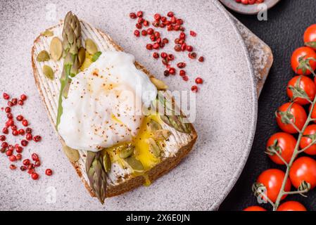 Colazione deliziosa e abbondante, con uova in camicia su pane tostato con formaggio spalmabile, asparagi, sale e spezie su sfondo di cemento scuro Foto Stock