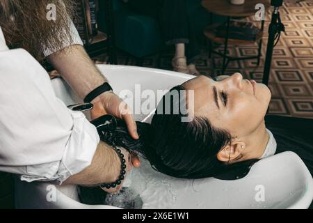 Parrucchiere che lava i capelli di una donna. Uomo giovane parrucchiere professionista esperto in gilet con barba lunga e capelli, capelli in lavaggio testa di capelli cliente femminile Foto Stock