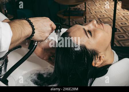 Parrucchiere che lava i capelli di una donna. Uomo giovane parrucchiere professionista esperto in gilet con barba lunga e capelli, capelli in lavaggio testa di capelli cliente femminile Foto Stock