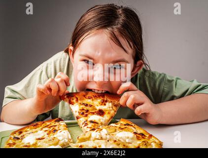 Una ragazza pre-adolescente mangia una fetta di pizza. Una bambina affamata con una t-shirt verde prende avidamente un boccone di una pizza grande. Primo piano, fuoco scelto. Foto di alta qualità Foto Stock