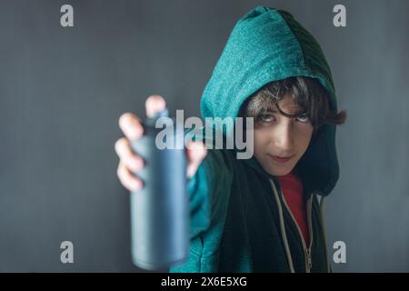 Un giovane con cappuccio verde ha in mano una lattina di vernice spray per graffiti. Guarda la telecamera con un'espressione seria. Foto Stock