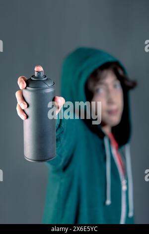 Un ragazzo tiene in mano una lattina di vernice spray. Indossa una felpa verde con cappuccio e un sorriso sul viso. Concetto di divertimento creativo con graffiti Foto Stock