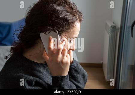 Primo piano di una donna seria che partecipa a una telefonata seduta su un letto in camera da letto Foto Stock