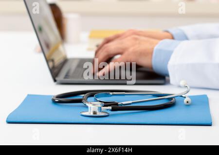 Primo piano di uno stetoscopio appoggiato su un computer portatile con un medico irriconoscibile che lavora in background con un computer portatile Foto Stock