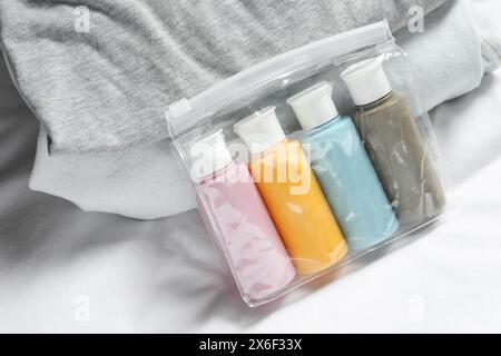 Kit da viaggio cosmetico. Sacchetto di plastica con piccoli contenitori di prodotti per la cura personale e pila di vestiti sul letto Foto Stock