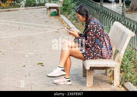 Una giovane donna con uno zaino è seduta su una panchina e guarda un telefono cellulare Foto Stock
