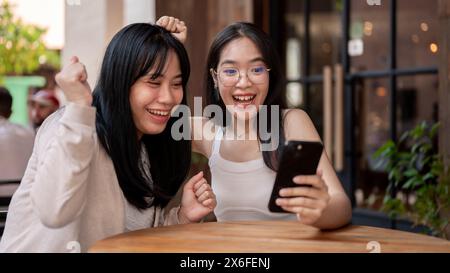 Due amiche asiatiche entusiaste e allegre guardano uno smartphone con volti estremamente felici, festeggiando insieme buone notizie mentre sono sedute Foto Stock