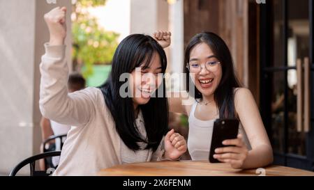 Due amiche asiatiche entusiaste e allegre guardano uno smartphone con volti estremamente felici, festeggiando insieme buone notizie mentre sono sedute Foto Stock