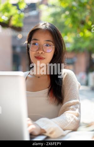 Una bella giovane donna asiatica con occhiali e abbigliamento casual si sta concentrando sul suo lavoro sul suo computer portatile, leggendo giornali online, pensando e pianificando Foto Stock