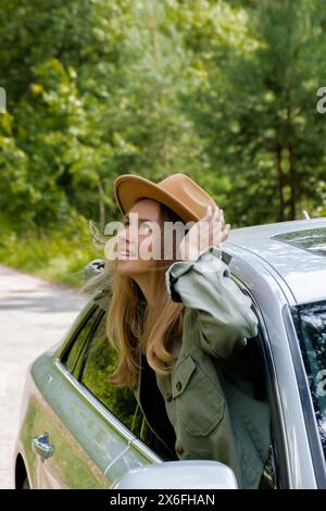 Donna bionda con cappello che sporge la testa dalla vettura del parabrezza. I giovani turisti esplorano i viaggi locali, trasformandoli in momenti autentici. Emozioni vere espressioni di fuga e di rinfrescarsi rilassarsi all'aria aperta e pulita Foto Stock