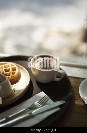 Caffè cappuccino e waffle con forchetta e coltello sullo sfondo bokeh alla luce del sole. Foto Stock