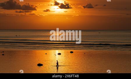 Il tramonto si aggira, il tramonto mozzafiato su Bining Beach, Bali, catturando la bellezza serena delle onde che si infrangono dolcemente contro la barriera corallina mentre la gente passeggia Foto Stock