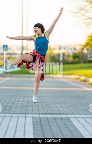 Una giovane donna gioiosa che indossa le cuffie balla giocosamente in un parco cittadino, vestita con una canotta blu e una camicia a quadri rossa legata alla vita. Foto Stock