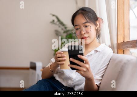 Una giovane donna asiatica che si rilassa a casa, legge il blog online sul suo smartphone mentre sorseggia un caffè sul divano in salotto. persone e stile di vita Foto Stock