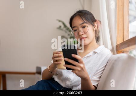 Una giovane donna asiatica che si rilassa a casa, legge il blog online sul suo smartphone mentre sorseggia un caffè sul divano in salotto. persone e stile di vita Foto Stock